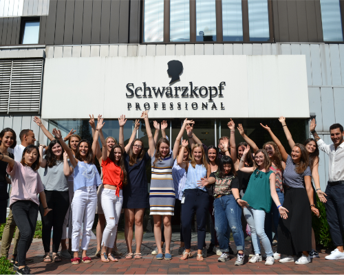 Un diverso equipo de Henkel de pie animando frente al edificio profesional de Schwarzkopf y levantando los brazos