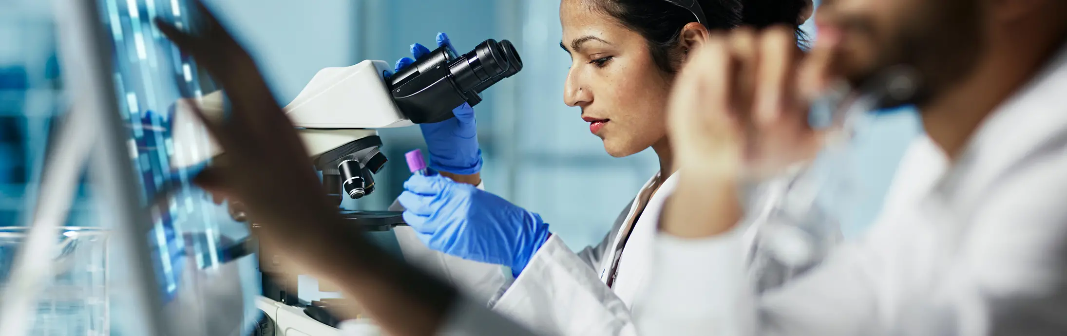 Un laboratorio con una mujer sentada delante de un microscopio, mirando una muestra y en frente un hombre con barba observando una pantalla. 