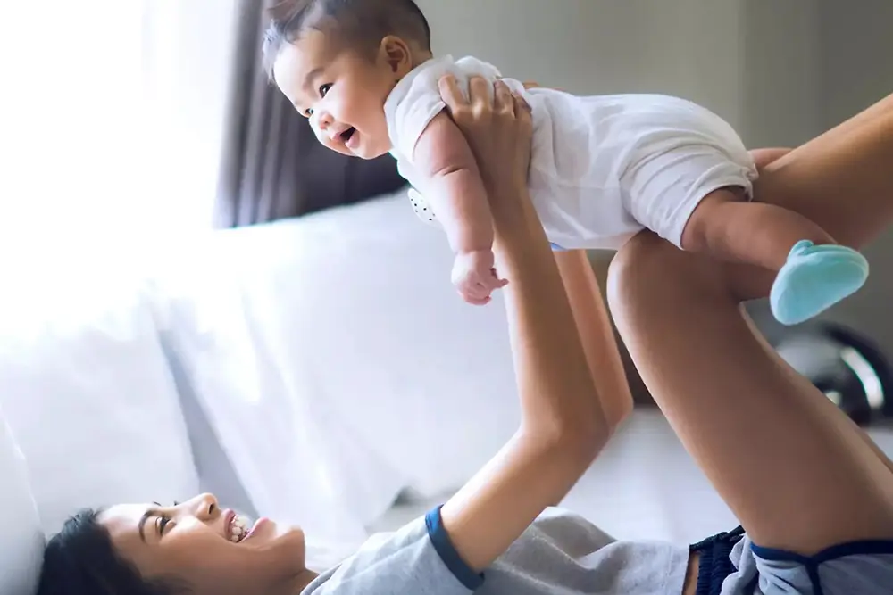 Una madre levanta y juega con un bebé sonriente en el interior de una casa luminosa.