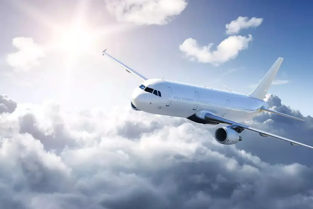 Un avión de pasajeros blanco volando sobre nubes suaves en un cielo brillante, con la luz del sol brillando desde la parte superior izquierda