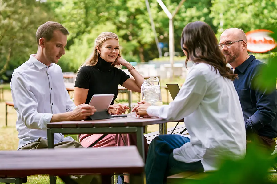 Grupo de colegas sentados juntos al aire libre, hablando sobre el trabajo con tabletas y computadoras portátiles, lo que representa la colaboración y una cultura abierta y orientada al equipo en Henkel.
