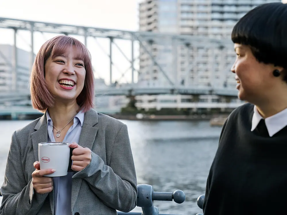 Two people having a casual conversation by a waterfront railing, one holding a Henkel-branded coffee mug, with a bridge and city buildings in the background—illustrating Henkel’s flexible work culture.