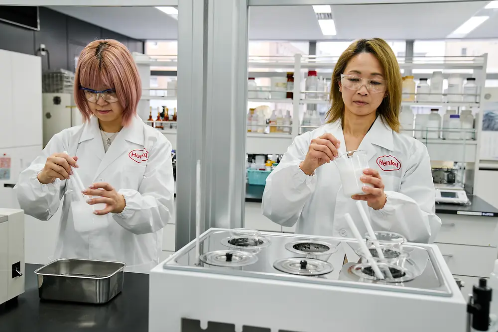 Dos personas con batas de laboratorio blancas de Henkel están trabajando en un laboratorio moderno. Están manipulando instrumentos científicos y recipientes en una mesa de trabajo, con varios equipos de laboratorio y estantes de suministros visibles al fondo.