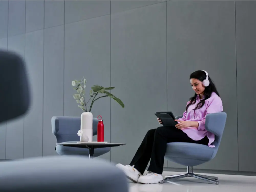 Una persona sentada en un sillón moderno, con audífonos y usando una tableta en un espacio de oficina minimalista. En una pequeña mesa redonda cercana hay un jarrón con plantas y una botella de agua roja. La habitación cuenta con paredes revestidas de paneles grises, iluminación tenue y muebles contemporáneos.