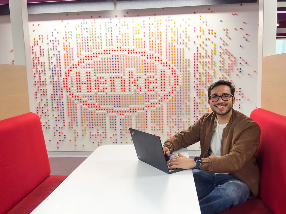Una persona sentada en una moderna cabina de oficina con un banco tapizado en rojo, trabajando en una laptop sobre una mesa blanca. Detrás se observa un exhibidor decorativo con pines de colores alrededor del logotipo rojo de Henkel.