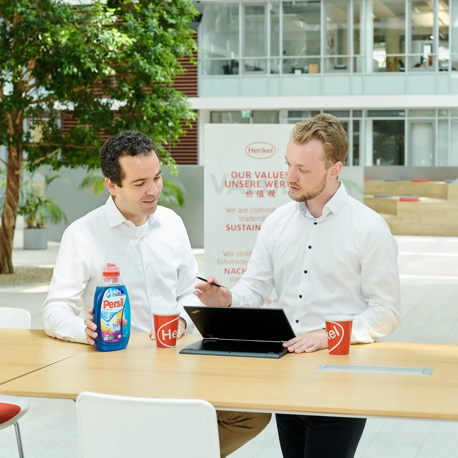 Dos hombres de pie en una mesa con una botella de Persil y una computadora portátil